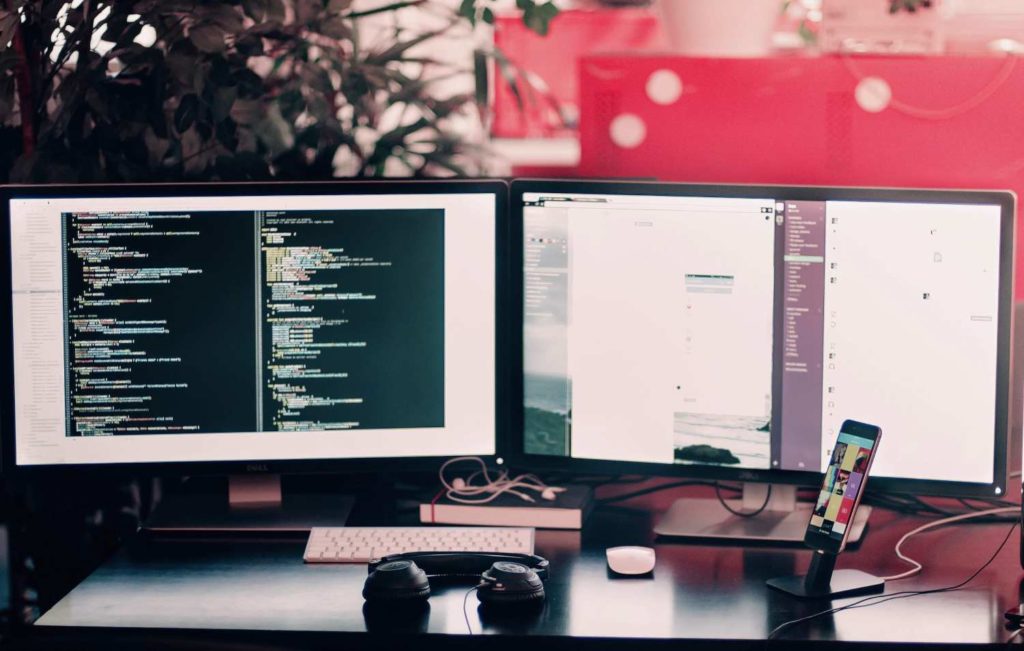 Two computer monitors sit side by side on a desktop with a mobile, reviewing website code