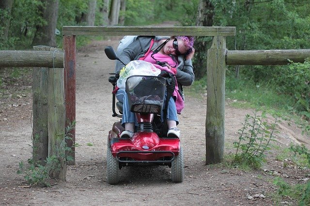 A girl with pink hair is using a red mobility scooter and ducking her head down, to try to gain access through a wooden fence which was not built for full disabled access.