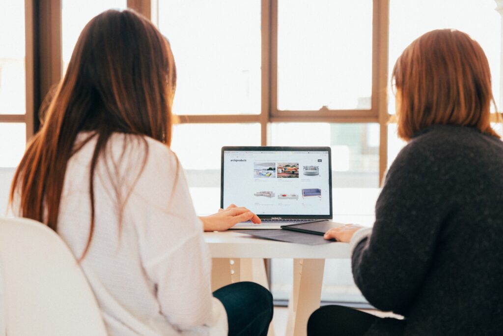 we are looking over the shoulders of two women sitting at a laptop which is open as they share resources.
