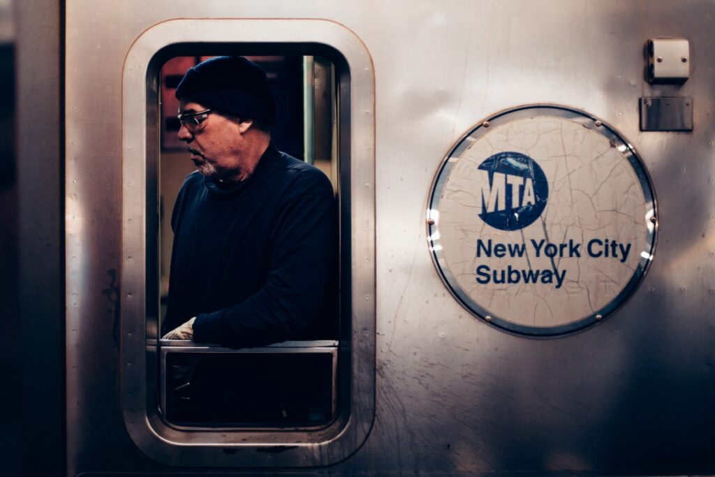 a middle aged man wearing black glasses and a beanie in a train window next to sign reading, New York City Subway.