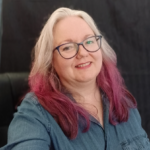 Suzy Molloy wearing blue framed glasses and looking towards the camera from an angle, Suzy has ombre platinum to pink to purple hair and is wearing a blue denim shirt.